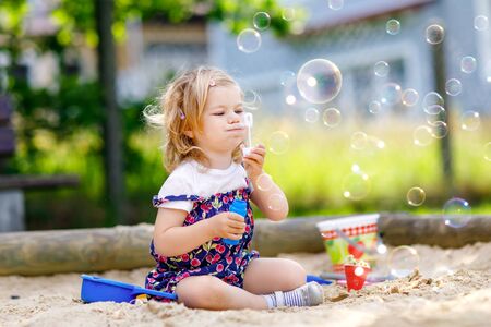 Beautiful little blonde toddler girl having fun with blowing soap bubble blower. Cute adorable baby child playing on playground on sunny summer day. Happy active funny healthy kidの写真素材
