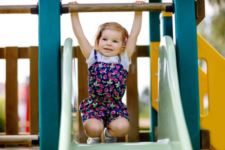 Cute toddler girl playing on slide on outdoor playground. Beautiful baby in colorful shorts trousers...