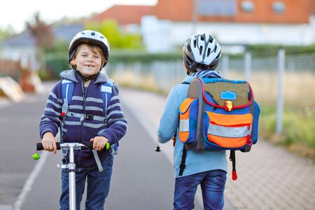 Two school kid boys in safety helmet riding with scooter in the city with backpack on sunny day. Happy children in colorful clothes biking on way to school.の写真素材
