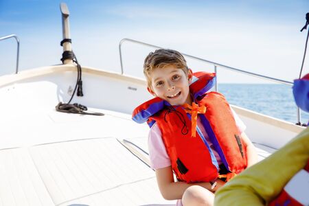 Happy blonde kid boy enjoying sailing boat trip. Family vacations on ocean or sea on sunny day. Healthy beautiful school child smiling and having fun on yacht. Coastline with villages and natureの写真素材