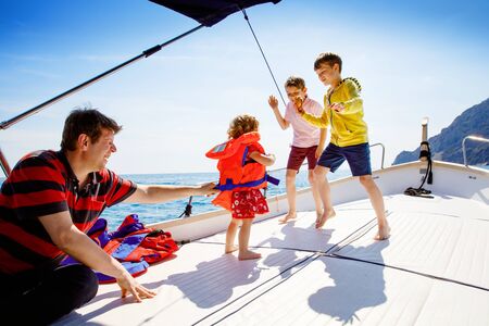 Two little kid boys, father and toddler girl enjoying sailing boat trip. Family vacations on ocean or sea on sunny day. Children smiling. Brothers and sister, siblings and dad having fun on yacht.の写真素材