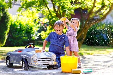 Two happy children washing big old toy car in summer garden, outdoors. Brother boy and little sister toddler girl cleaning car with soap and water, having fun with splashing and playing with sponge.の写真素材