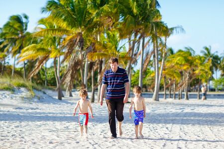Two happy little kids boys and father walking on the beach of ocean with palms on sunny day. Family, dad and cute children sons making vacations, dreaming and enjoying summerの写真素材