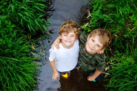 Two happy little school kids boys, funny siblings having fun together walking through water in river in gum rubber boots. Family portrait of healthy brothers and best friendsの写真素材