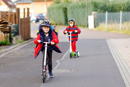 Two little kids boys riding on push scooters on the way to or from school. Schoolboys of 7 years driving through rain puddle. Funny siblings and best friends playing together. Children after schoolの写真素材