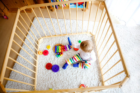 Beautiful little baby girl standing inside playpen. Cute adorable child playing with colorful toys....