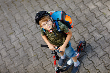 Two school kid boys in safety helmet riding with scooter in the city with backpack on sunny day....