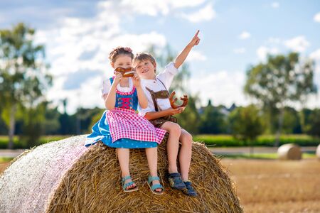 Two kids in traditional Bavarian costumes in wheat field. German children eating bread and pretzel during Oktoberfest. Boy and girl play at hay bales during summer harvest time in Germany.の写真素材