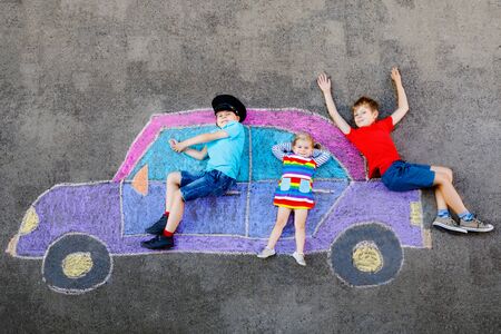 three little children, two school kids boys and toddler girl having fun with with car picture drawing with colorful chalks on asphalt. Siblings painting on ground playing together. Creative leisureの写真素材
