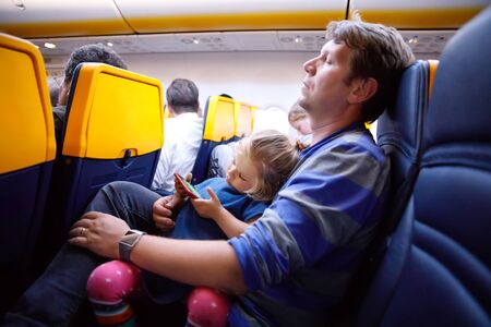 Young father holding his baby toddler daughter during flight on airplane going on vacations. Tired man and cute girl during night flight. child playing with smartphone. Family sitting in planeの写真素材