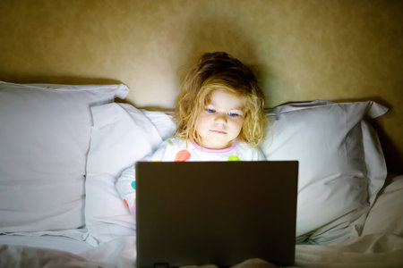 Cute adorable little toddler girl watching cartoons on laptop in bed. Tired baby child with tablet...