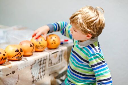Little kid boy making Jack-o-lantern with drawing scary faces on mandarine, tangerine or clementine. Happy child making preparation for Halloween party at home, indoorsの写真素材
