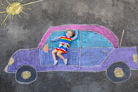 Adorable little toddler girl playing with colorful chalks and painting big car picture on asphalt. Happy baby child playing outside. Creative leisure for children outdoors in summerの写真素材