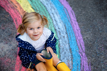 Happy little toddler girl in rubber boots with rainbow sun and clouds with rain painted with...