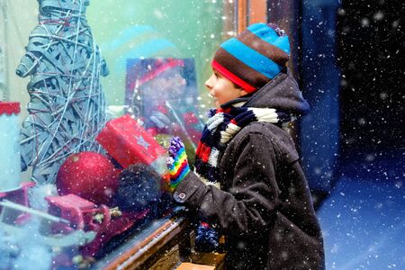 Cute little healthy school kid boy on Christmas market. Funny happy child in fashion winter clothes making window shopping decorated with gifts, xmas tree. Snow falling down, snowfallの写真素材
