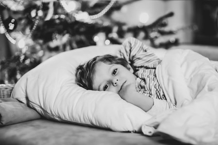 Little cute blond boy sleeping under Christmas tree and dreaming of Santa at home, indoors. Traditional Christian festival. Happy kid child waiting for gifts on xmas. Cozy soft lightの写真素材