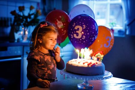 Adorable little toddler girl celebrating third birthday. Baby toddler child with homemade unicorn cake, indoor. Happy healthy toddler is suprised about firework sparkler and blowing candles on cakeの写真素材