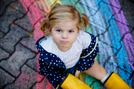 Happy little toddler girl in rubber boots with rainbow sun and clouds with rain painted with...