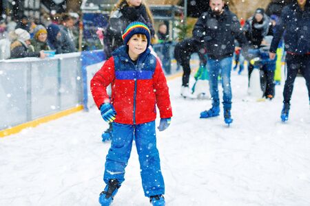 Happy little kid boy in colorful warm clothes skating on a rink of Christmas market or fair. Healthy child having fun on ice skate. Lot of people celebrating holiday and having active winter leisureの写真素材