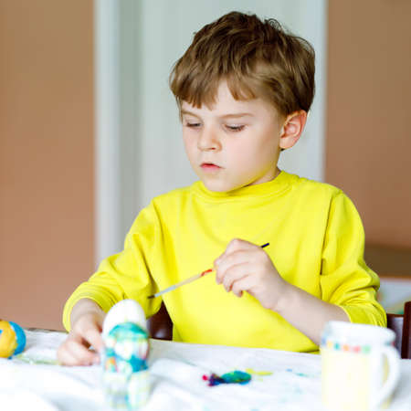 Little blond preschool kid boy coloring eggs for Easter holiday in domestic kitchen, indoors. Child having fun with painting colors and celebrating feast.の写真素材