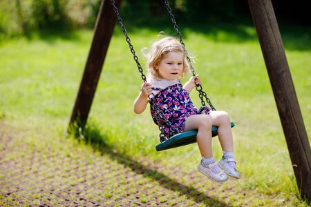 Cute adorable toddler girl swinging on outdoor playground. Happy smiling baby child sitting in chain swing. Active baby on sunny summer day outsideの写真素材