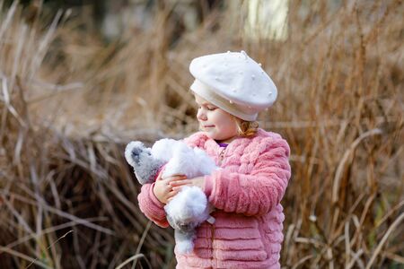 Outdoor portrait of little cute toddler girl in pink coat and white fashion hat barret playing with soft plush dog toy. Healthy happy baby child walking in the park on cold day.の写真素材