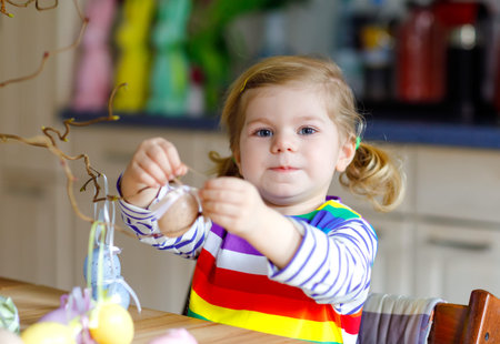 Cute little toddler girl decorating tree bough with colored pastel plastic eggs. Happy baby child...