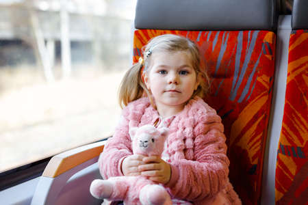 Cute little toddler girl sitting in train and looking out of window while moving. Adorable happy healthy baby child holding plush toy in hands. Smiling child going on family vacations by railroad.の写真素材