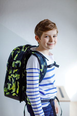 Beautiful kid boy with schoolbag rucksack standing in room in early morning. Happy healthy child on...