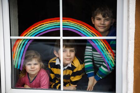 Three kids, two school kids boys and toddler girl with rainbow painted with colorful window color during pandemic coronavirus quarantine. Children painting rainbows with the words Lets all be well.の写真素材