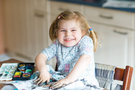 Cute adorable baby girl learning painting with water colors. Little toddler drawing at home, using colorful brushes. Healthy happy child with dirty face and hands at home or nurseryの写真素材