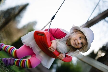 Cute adorable toddler girl swinging in domestic garden with soft plush toy. Happy healthy baby child in hat and dress playing outdoors on sunny warm day.の写真素材