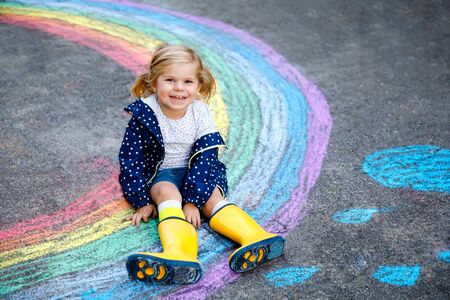 Happy little toddler girl in rubber boots with rainbow painted with colorful chalks on ground during pandemic coronavirus quarantine. Children painting rainbows along with the words Lets all be wellの写真素材