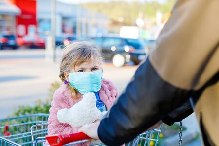Toddler girl in medical mask as protection against pandemic coronavirus disease. Cute child using protective equipment against covid 19 and shopping in supermarket with cart pushing by fatherの写真素材