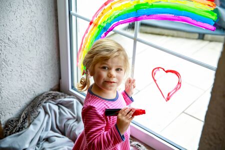 Adoralbe little toddler girl with rainbow painted with colorful window color during pandemic coronavirus quarantine. Child painting rainbows around the world with the words Lets all be well.の写真素材