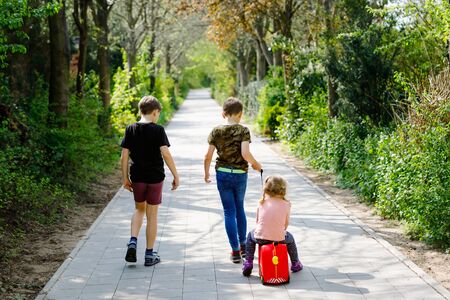 three kids, little toddler girl and two kid boys walking in park pandemic coronavirus disease. Children, lovely siblings playing together as family, using toy car suitcase on wheelsの写真素材