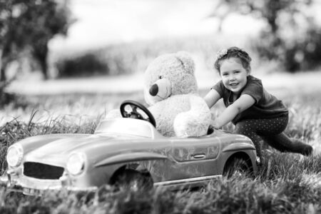 Little preschool kid girl driving big vintage old toy car and having fun with playing with big plush toy bear, outdoors. Child enjoying warm summer day in nature landscape. Girl driving carの写真素材