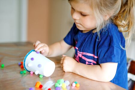 little toddler girl making craft lantern with paper cups, colorful pompoms and glue during pandemic coronavirus quarantine disease. Happy creative child, homeschooling and home daycare with parentsの写真素材