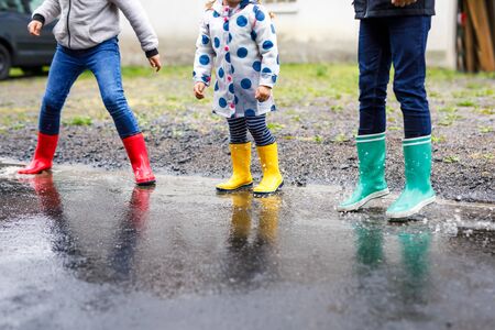 Close-up of three children, toddler girl and two kids boys wearing red, yellow and green rain boots and walking during sleet. Happy siblings jumping into puddle. Having fun outdoors, active family.の写真素材