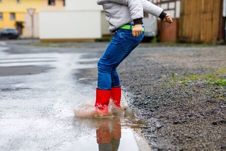 Close-up of kid boy wearing red rain boots and walking during sleet and rain on rainy cloudy day. Child in colorful casual clothes jumping into puddle. Having fun outdoors, healthy children activity.の写真素材