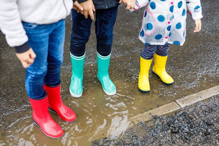 Close-up of three children, toddler girl and two kids boys wearing red, yellow and green rain boots and walking during sleet. Happy siblings jumping into puddle. Having fun outdoors, active familyの写真素材