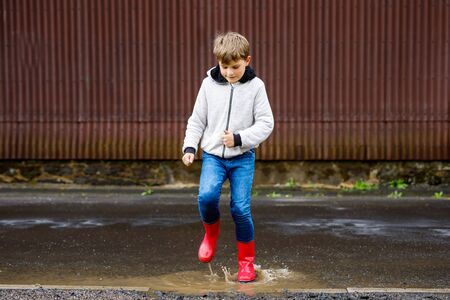 Happy kid boy wearing red rain boots and walking during sleet and rain on rainy cloudy day. Child in colorful casual clothes jumping into puddle. Having fun outdoors, healthy children activityの写真素材