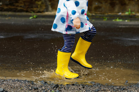 Little toddler girl wearing yellow rain boots, running and walking during sleet on rainy cloudy day. Cute happy child in colorful clothes jumping into puddle, splashing with water, outdoor activityの写真素材
