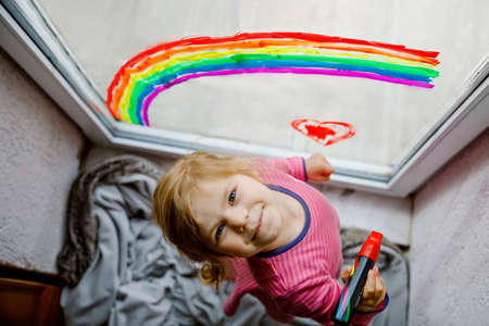 Adoralbe little toddler girl with rainbow painted with colorful window color during pandemic coronavirus quarantine. Child painting rainbows and hearts around the world with words Lets all be well.の写真素材