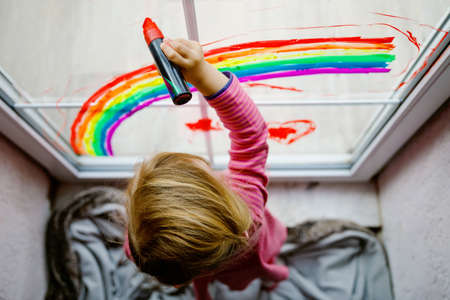 Adoralbe little toddler girl with rainbow painted with colorful window color during pandemic coronavirus quarantine. Child painting rainbows and hearts around the world with words Lets all be well.の写真素材