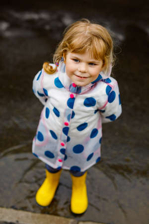 Little toddler girl wearing yellow rain boots, running and walking during sleet on rainy cloudy day. Cute happy child in colorful clothes jumping into puddle, splashing with water, outdoor activityの写真素材