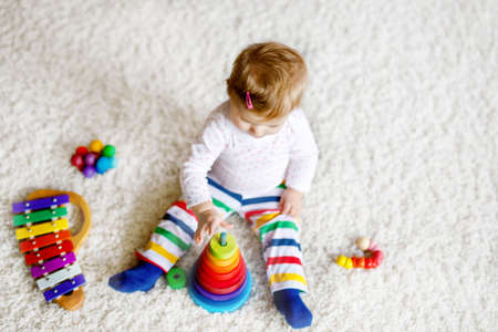 Adorable cute beautiful little baby girl playing with educational toys at home or nursery. Happy healthy child having fun with colorful wooden rainboy toy pyramid. Kid learning different skillsの写真素材