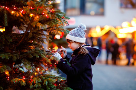 Little cute kid girl having fun on traditional Christmas market during strong snowfall. Happy child enjoying traditional family market in Germany. Schoolgirl standing by illuminated xmas treeの写真素材