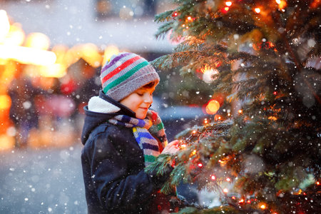 Little cute kid boy having fun on traditional Christmas market during strong snowfall. Happy child enjoying traditional family market in Germany. Schoolboy standing by illuminated xmas treeの写真素材