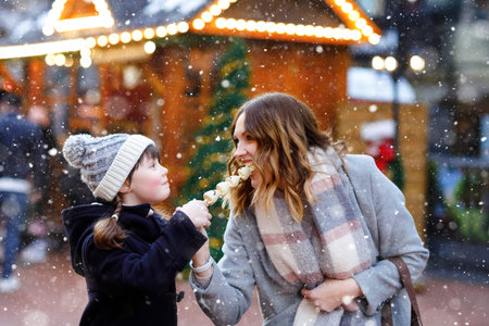 Young mother and daughter eating white chocolate covered fruits and strawberry on skewer on traditional German Christmas market. Happy girl and woman on traditional family market in Germany during snowy dayの写真素材